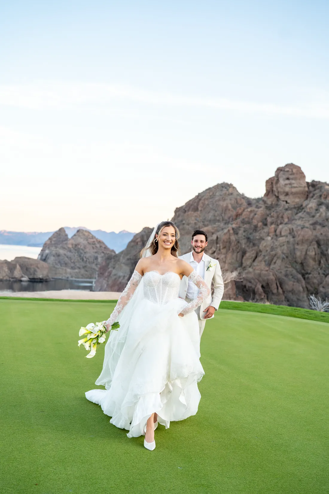 La novia camina por el green levantando el vestido, con las rocas atrás
