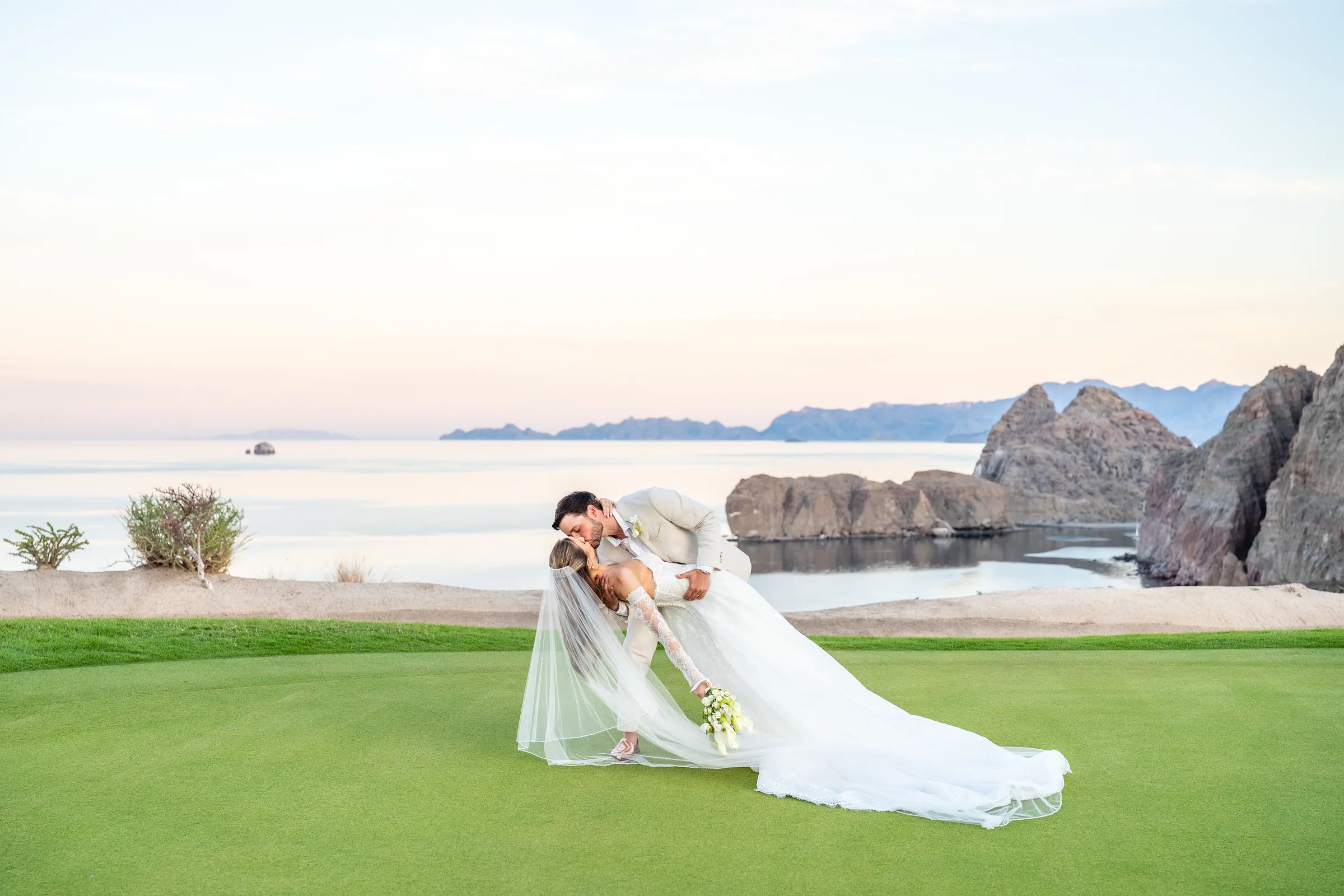 Hollye y Maccoy se besan sobre el green del campo de golf, con el velo extendido y el Mar de Cortés al atardecer, en Villa del Palmar, Loreto