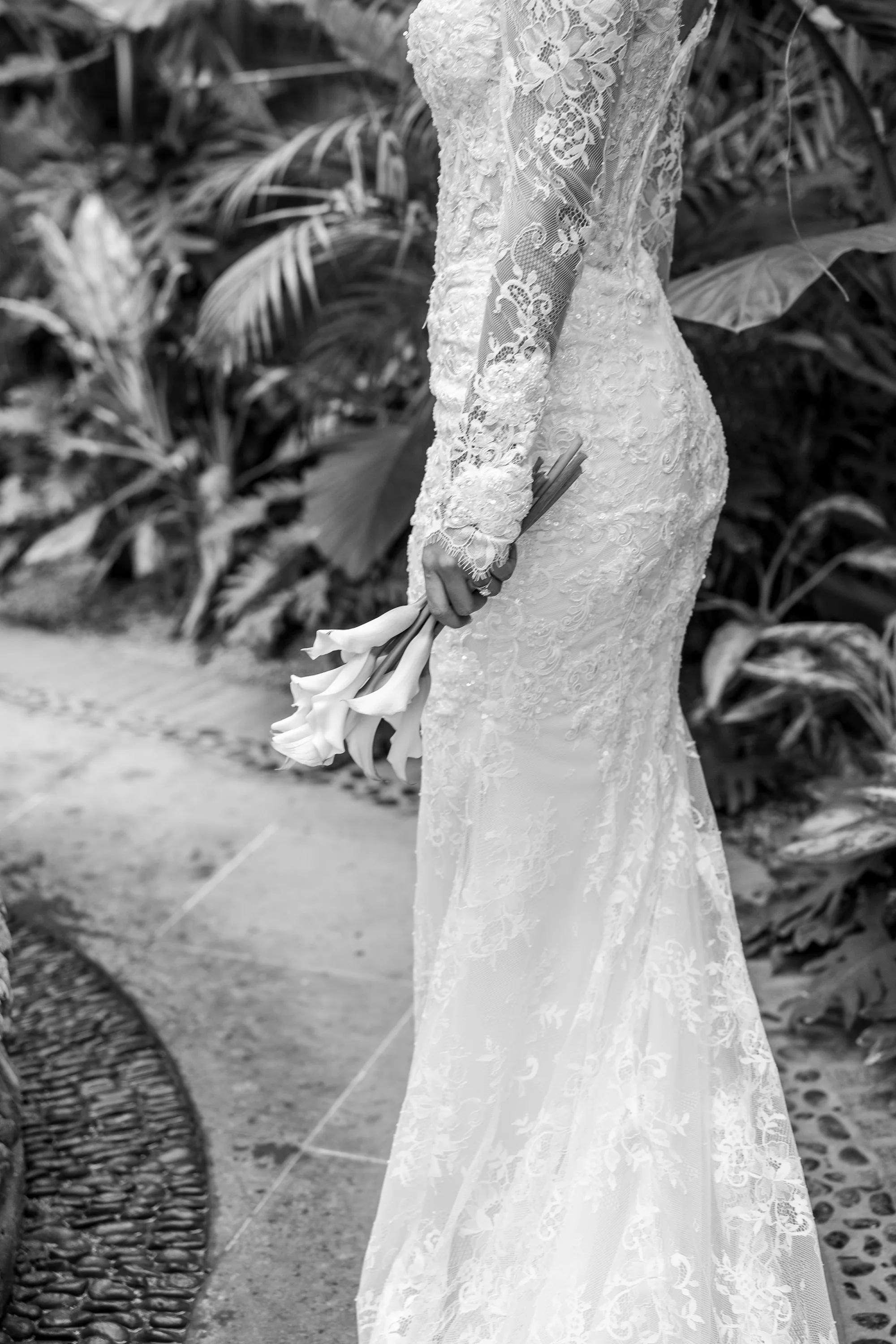 Detalle en blanco y negro del encaje del vestido de novia y el ramo de alcatraces, entre la vegetación tropical