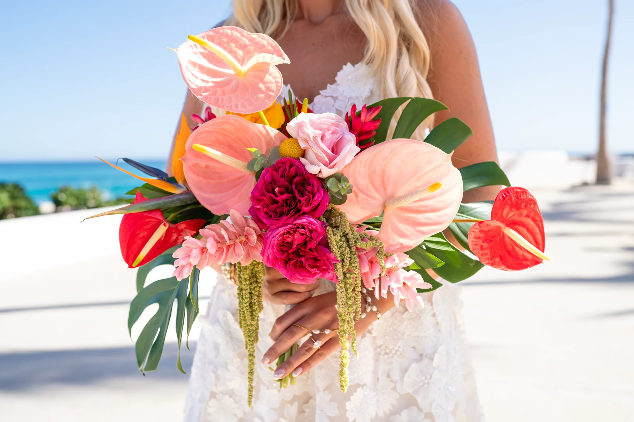 La novia sostiene un ramo tropical de anturios, rosas y hojas de monstera frente al mar