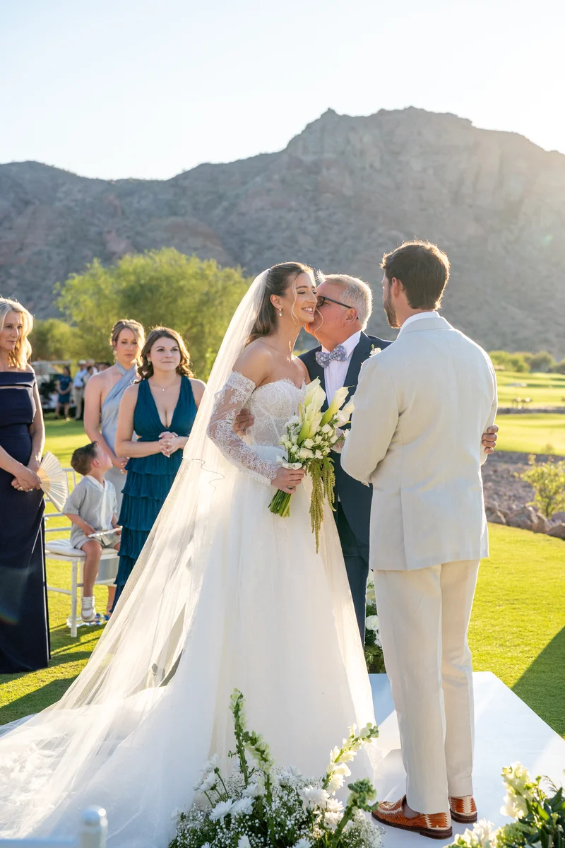El padre de la novia la besa en la mejilla frente al altar mientras el novio los observa