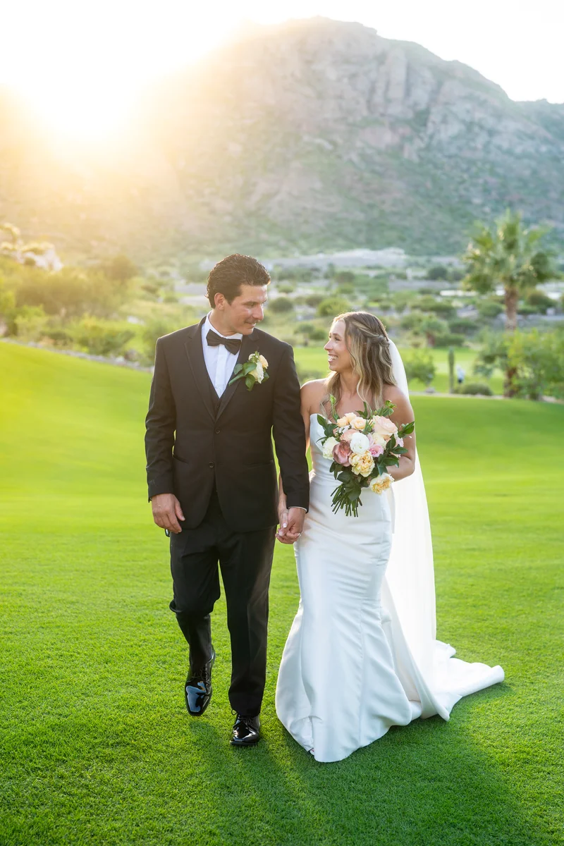 Los novios caminan tomados de la mano sobre el pasto con la montaña al atardecer