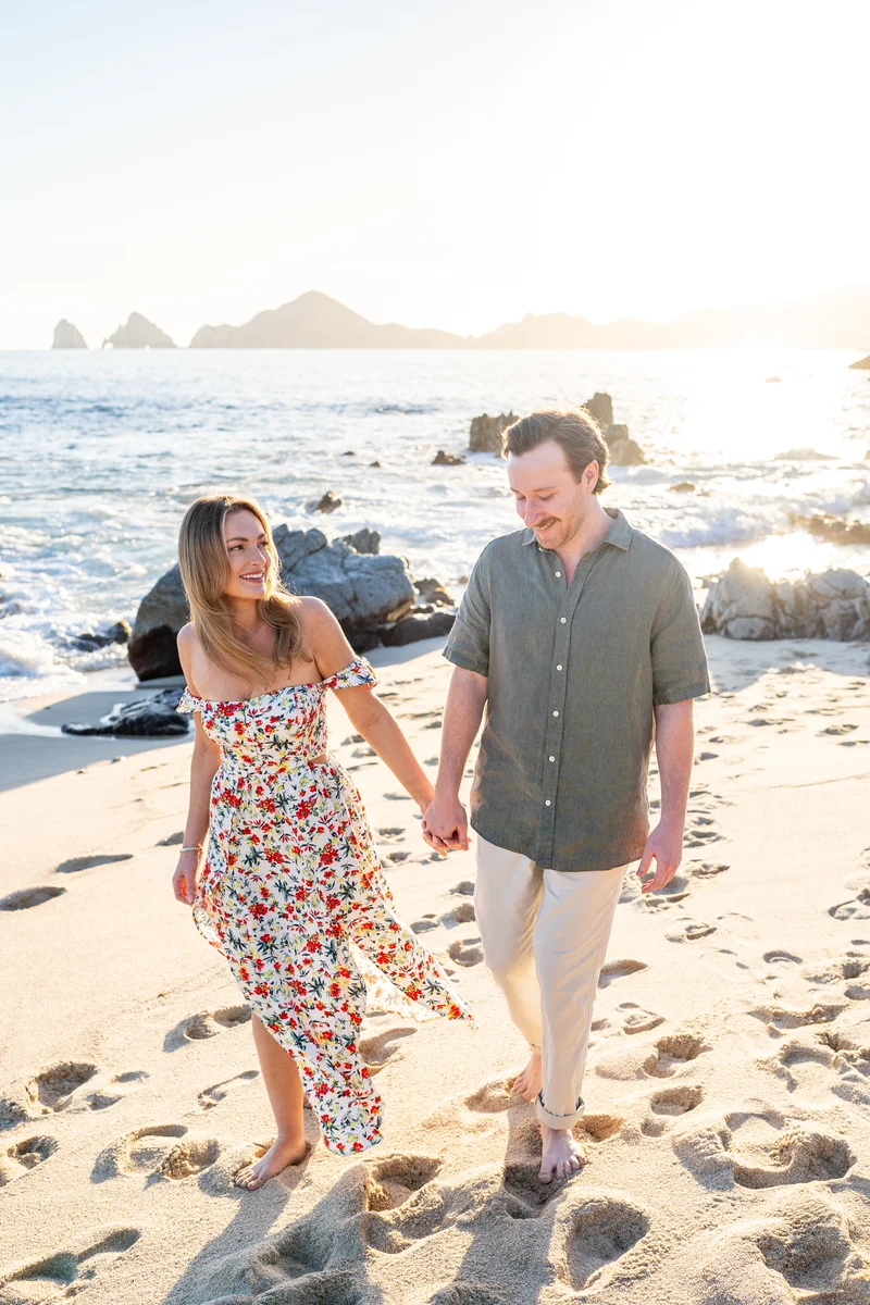 Pareja caminando de la mano por la playa de Cabo San Lucas con El Arco al fondo